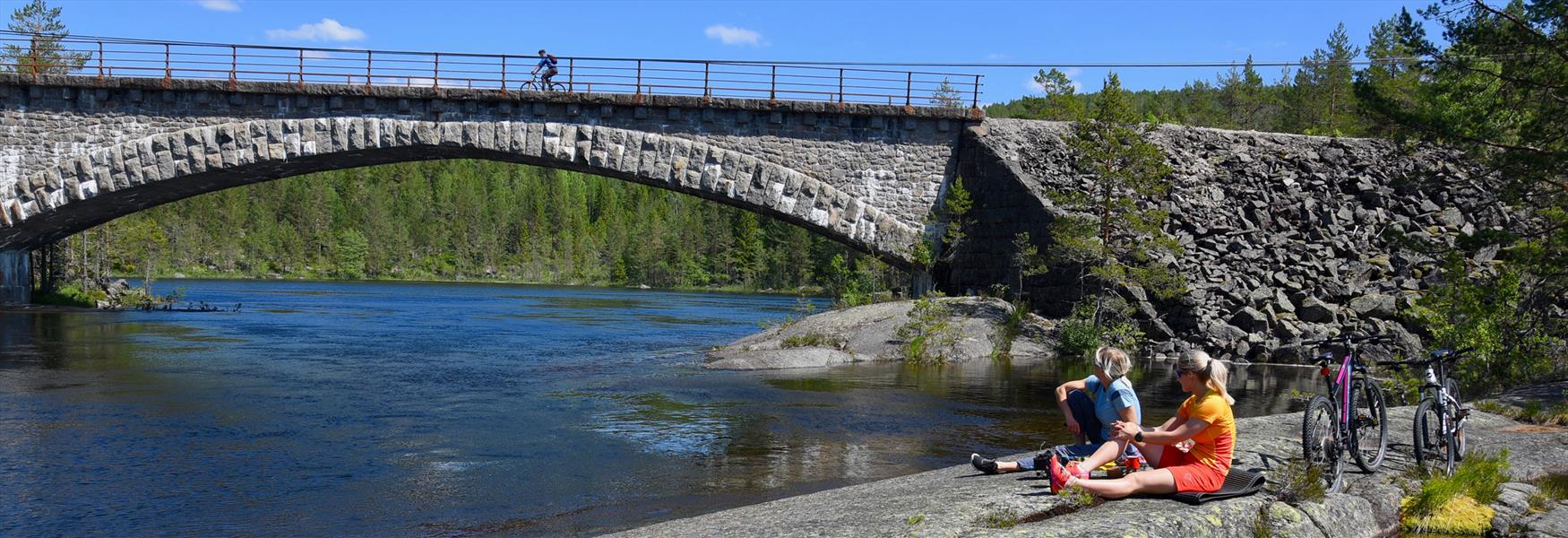 2 damer med sykler sitter på en stein ved en elv og se på syklisten sykler over bro