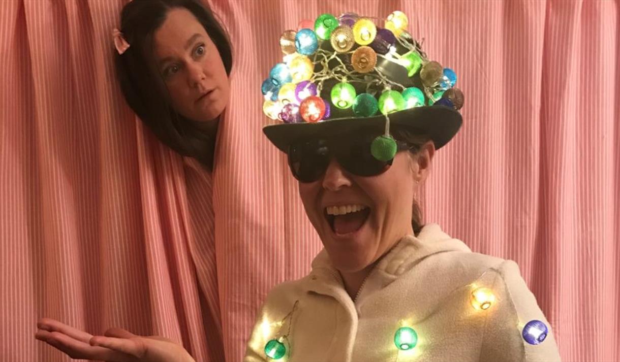 En dame med hatt med lyslenker på og lyslenker hengende på seg. En annen dame stikker hode ut fra bak en gardin