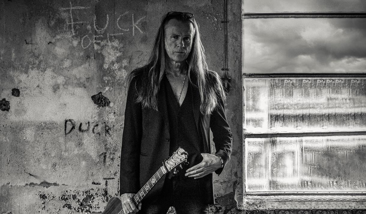 mann med langt blondt hår , sort jakke og gitar,