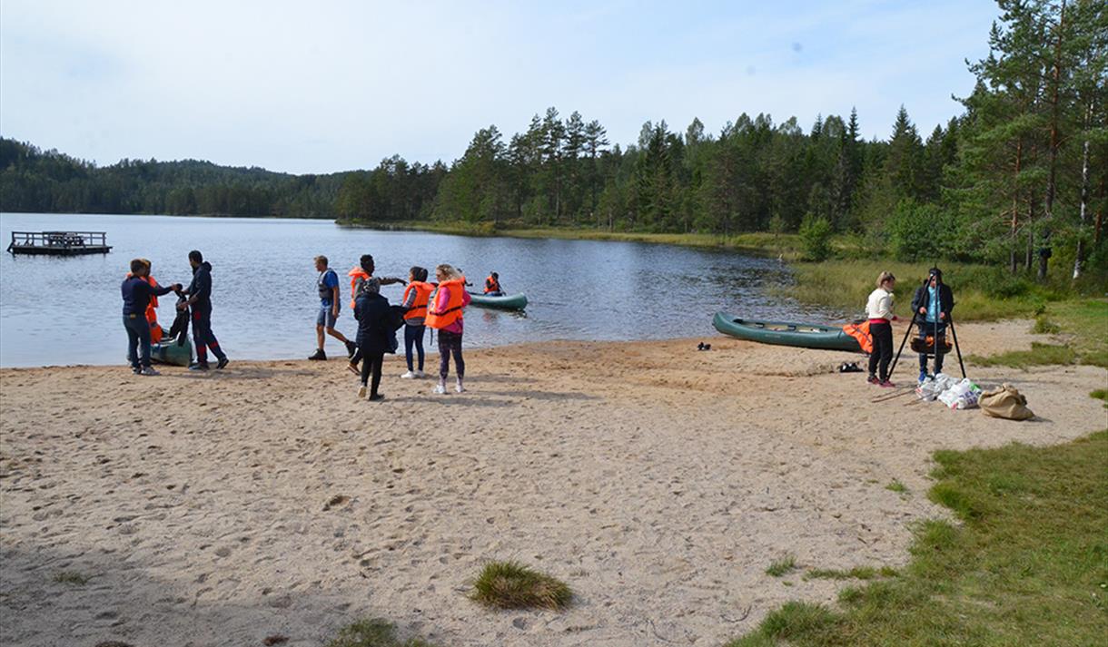 sandstrand med kano og ungdommer, flytebrygge og skogsområder