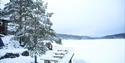 Brygge med bord, snø og grantrær, hytte i bakgrunnen