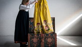 En dame i Telemarksbunad holder opp et skjørt i gult med blomster som har ligget i en rosemalt kiste