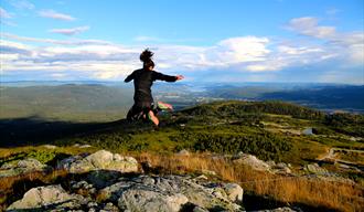 Photo from one of Lifjell's many summits. Blue sky, beautiful view of lake Norsjø and Telemark, and a girl jumping high in the air.