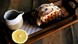 Cooked lobster served on a black tray on a wooden board, topped with herbs. A small jug of sauce and a halved lemon sit beside it on a rustic wooden t