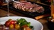 Close-up of sliced steak served on a hot rectangular platter, with tongs lifting a piece. A plate of salad with cherry tomatoes and greens sits blurre
