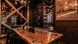 Warmly lit restaurant interior with a long wooden dining table set with wine glasses, cutlery, and a bottle of wine. Industrial-style décor features e
