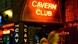 The exterior and entrance of the Cavern Club. The neon red entry sign reads 'Cavern Club.' There are two women leaving the club sminling.