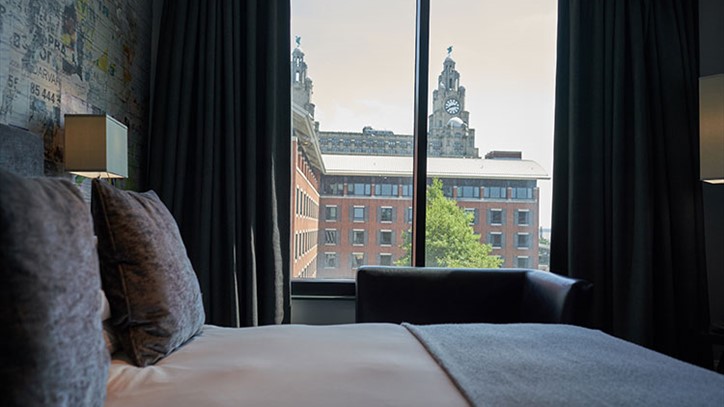 A large double bed with a cream cover and grey pillows with a window to the left with the Liver Building in view
