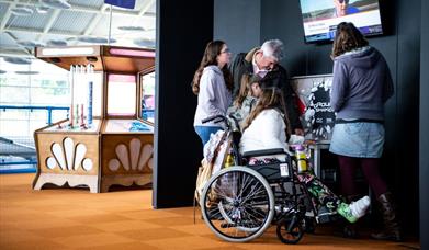 A family, featuring a girl in a wheelchair with a cast on her leg, and her parents and sibling looking at displays at Winchester Science Centre and Pl