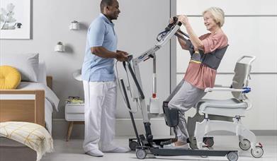 a carer helping a woman stand up in a chair using mobility hire equipment