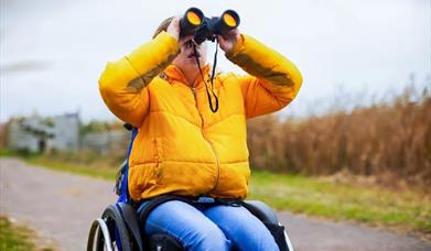 A child in a yellow coat and sat in a wheelchair with binoculars at WWT Arundel Wetland Centre