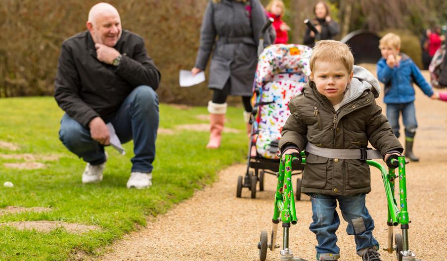 Little boy using a mobility aid to follow the path Sheffield Park and Gardens with other children and families in the background