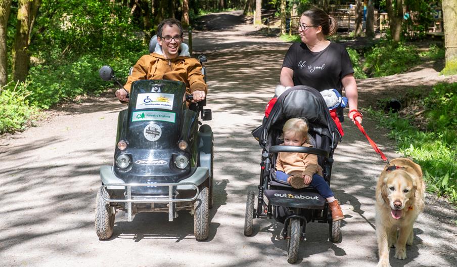 A family walking around a pathway at Wyre Forest, one on foot pushing a wheelchair and walking a dog and another on a tramper