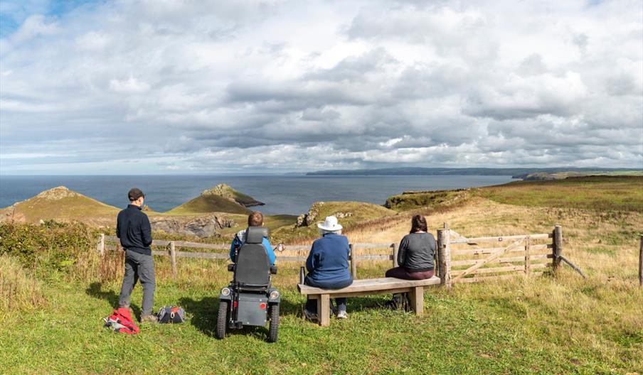 A group of people enjoying the view at National Trust Pentire, one on foot, one in an all terrain wheelchair and two sat on a bench