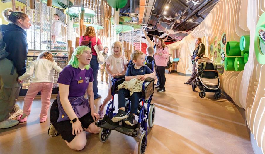 A woman is kneeled down with a boy in a wheelchair looking at a wooden display wall with other children in the background.