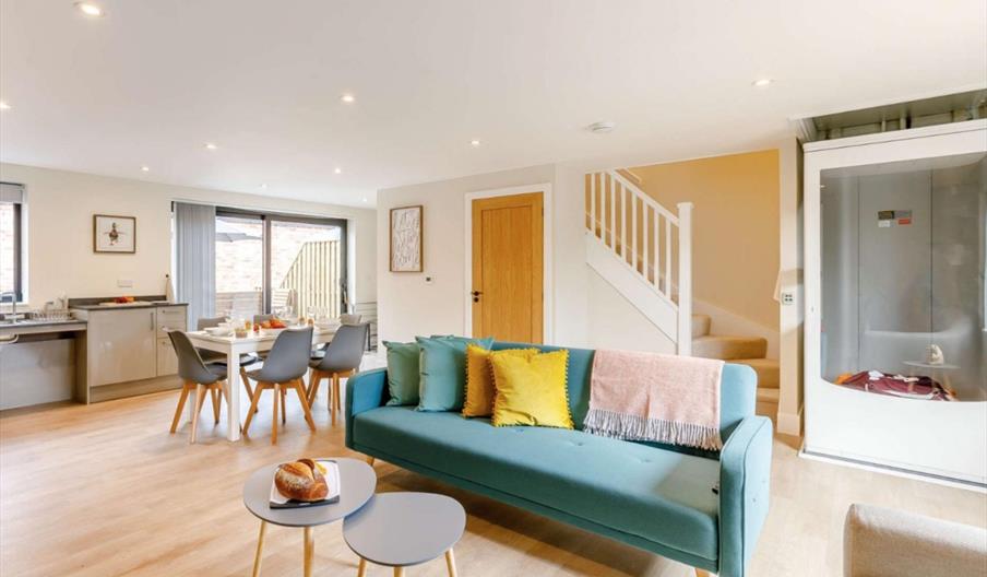 A modern open-plan living area with light wood flooring. Features a teal sofa with colorful cushions, dining set, stairs, and large window for natural
