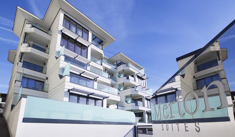 Modern apartment building under a clear blue sky, featuring white facades, large glass balconies, and a "Suites" sign conveying a luxury feel.