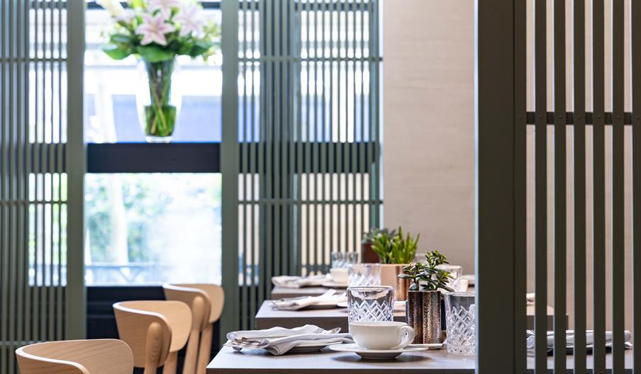 Stylish dining area with wooden furniture, set tables, crystal glasses, and a small plant. A vase of lilies decorates the window with vertical blinds.