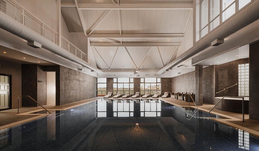 Spacious indoor swimming pool with sleek design, surrounded by lounge chairs. High ceiling and large windows create a calm, luxurious atmosphere.