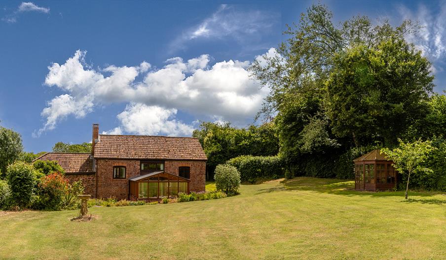 A charming brick cottage with a tiled roof sits amid lush greenery under a bright blue sky. A wooden gazebo stands to the right on a spacious lawn.