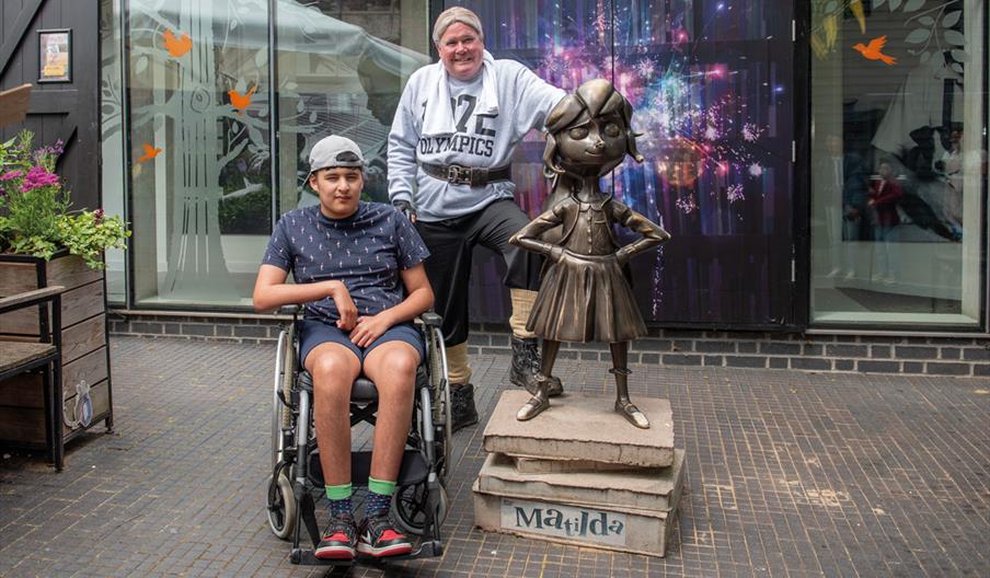 A young person in a wheelchair and an older person stand beside a bronze statue of Matilda. They are smiling, with a colorful, vibrant backdrop.
