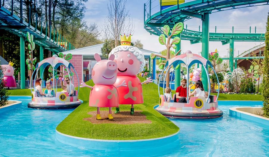 Colorful amusement park scene featuring large peppa pig statues on grassy island beside water ride with families in round boats. Playful, cheerful atm