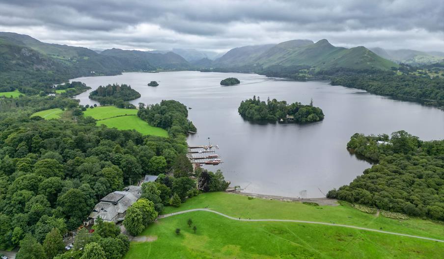 Aerial view of Derwentwater in the Lake District, featuring lush green landscapes, small forested islands, and boats by the shore under a cloudy sky.
