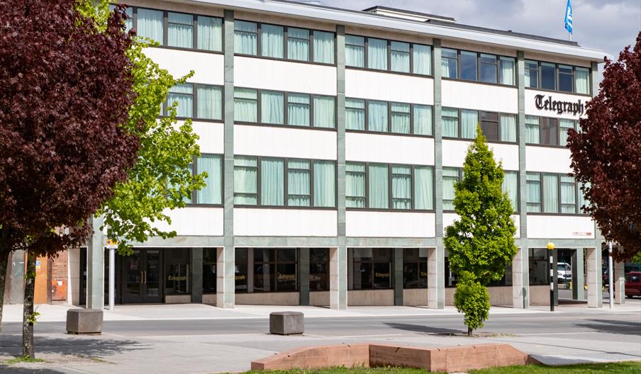 Modern office building labeled "Telegraph" with large grid windows, bordered by vibrant green and red trees, under a partly cloudy sky.