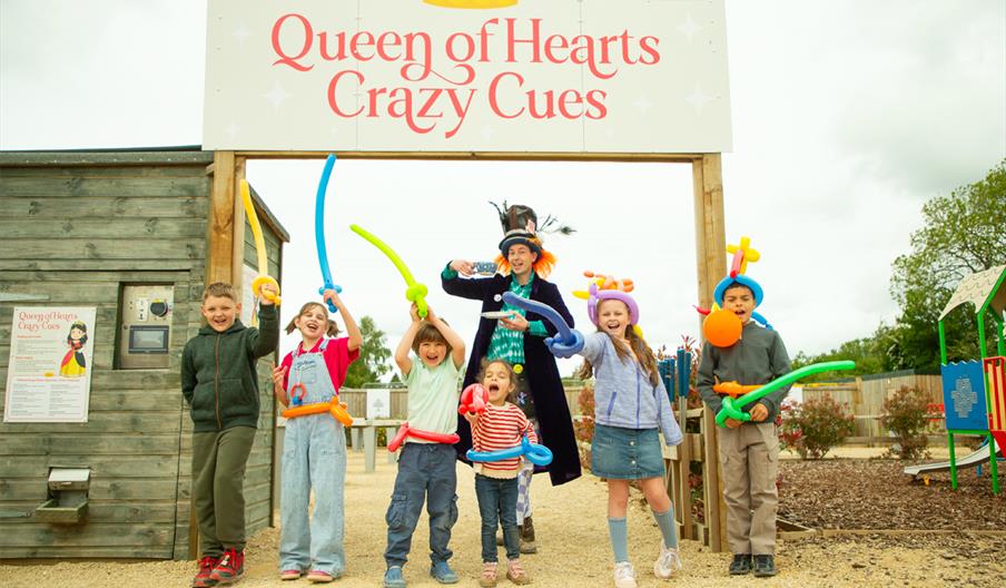 Six children with balloon swords stand in front of a sign reading 'Queen of Hearts Crazy Cues' with the Mad Hatter
