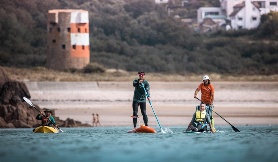 A group of people stand up paddleboarding, one supporting a disabled visitor with Healing Waves