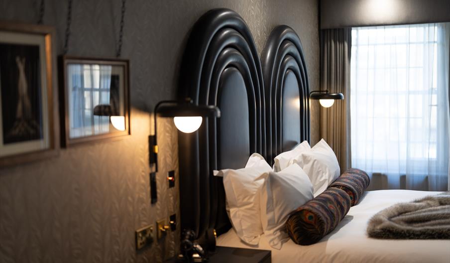 Luxurious hotel room with a double bed, ornate black headboard, plush pillows, and a textured blanket. Warm lighting and a large window create a cosy