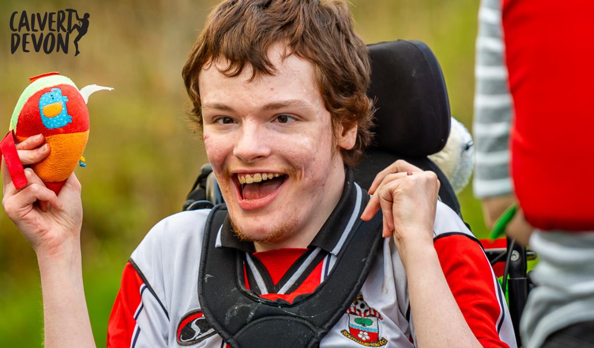 A joyful young person in a wheelchair holds a colorful plush toy, smiling widely outdoors. The bright and cheerful scene conveys happiness and excitem
