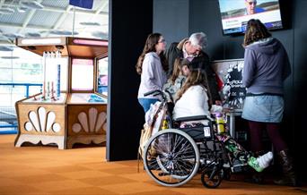 A family, featuring a girl in a wheelchair with a cast on her leg, and her parents and sibling looking at displays at Winchester Science Centre and Pl