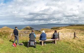 A group of people enjoying the view at National Trust Pentire, one on foot, one in an all terrain wheelchair and two sat on a bench