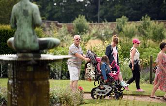 A group of people is walking through a lush garden with various plants and flowers. In the foreground, there is a bronze fountain with a statue of a s