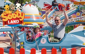 A parent and child on a plane ride at Crealy Theme Park with a Sooty Land logo over the top left corner