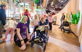 A woman is kneeled down with a boy in a wheelchair looking at a wooden display wall with other children in the background.