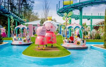 Colorful amusement park scene featuring large peppa pig statues on grassy island beside water ride with families in round boats. Playful, cheerful atm