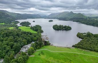 Aerial view of Derwentwater in the Lake District, featuring lush green landscapes, small forested islands, and boats by the shore under a cloudy sky.