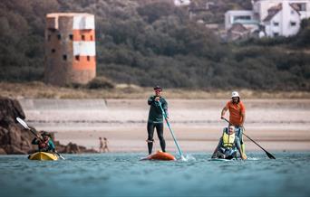 A group of people stand up paddleboarding, one supporting a disabled visitor with Healing Waves