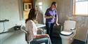 A guest being shown the accessible bathroom at Hoe Grange Holidays, there are handles either side of the toilet and a shower chair