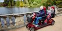 Two people riding an all terrain mobility scooter with two seats at Sheffield Park and Gardens