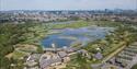 Aerial view of a lush wetland reserve with winding ponds and greenery. Buildings and a parking lot are in the foreground, with a cityscape in the dist