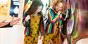
Two young children, both wearing bright yellow overalls with bee prints and rainbow-striped jackets, are engaged in an interactive exhibit. The child