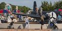 Children and adults enjoy a playground with airplane-themed structures, strollers, and benches in an outdoor setting.