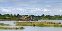 A serene lakeside scene with small islands covered in greenery and purple flowers. A building is visible in the background under a partly cloudy sky.