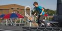 A boy in a blue shirt balances on a play structure in a playground with red and blue umbrellas in the background.