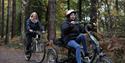 People riding bikes through the trees at Alice Holt Forest one using an adapted bike and one a standard bike