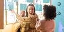 
A woman holding a baby is engaging with a young child in a playful, educational setting. She has a joyful, animated expression, pointing towards a wa