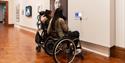 Two people in wheelchairs using the audio guides next to portraits at the National Portrait Gallery